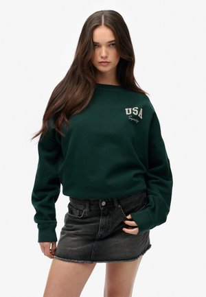 Sweat à capuche vert avec "USA" et "Superdry" en blanc, coupe décontractée, manches côtelées, associé à une mini-jupe en denim noir avec un ourlet effiloché.