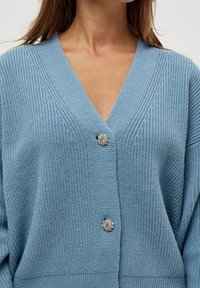 Cardigan lavorato a maglia blu chiaro con scollatura a V profonda, maniche ampie e un bottone decorativo a forma di fiore sul davanti.