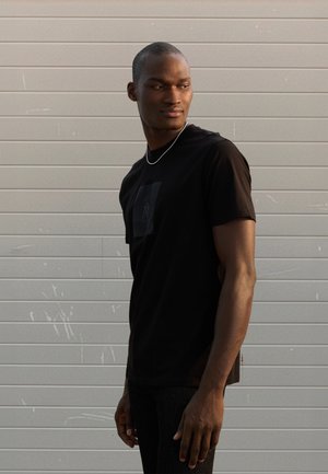 Black short-sleeved t-shirt with a square pocket, featuring a subtle logo print. Smooth fabric and relaxed fit, worn with dark jeans.
