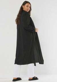 Donna con lunghi capelli castani, che indossa un lungo cappotto nero, pantaloni bianchi, sandali neri con borchie e orecchini a cerchio dorati, in piedi contro un muro bianco semplice.