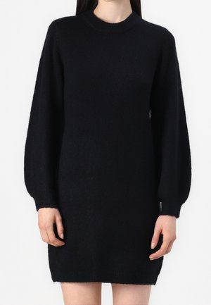 Femme portant une robe-pull noire à manches longues avec un col rond, debout devant un fond blanc uni.