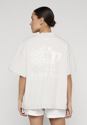 Kvinde iført hvid oversized t-shirt med tryk på ryggen af en kaffemaskine, blomster og teksten "de små ting" stående mod en ensfarvet baggrund.