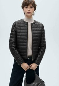 Jachetă puffer neagră cu un design matlasat, guler rotund și închidere cu fermoar. Purtați peste un pulover bej și asortată cu blugi întunecați.