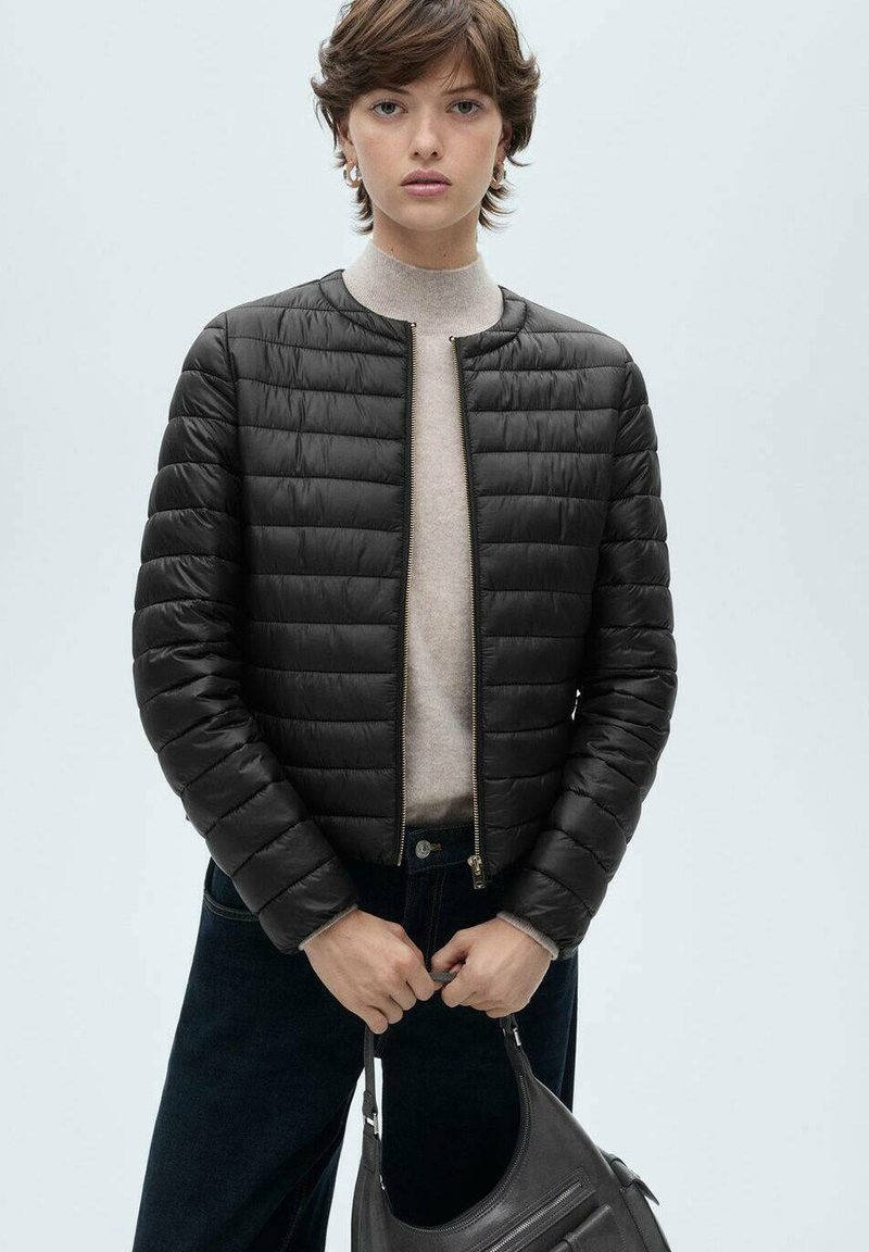 Jachetă puffer neagră cu un design matlasat, guler rotund și închidere cu fermoar. Purtați peste un pulover bej și asortată cu blugi întunecați.