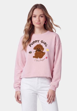 Giovane donna con capelli lunghi e mossi indossa una felpa rosa con "Puppy Girl" e una grafica di un cucciolo marrone, abbinata a pantaloni bianchi.