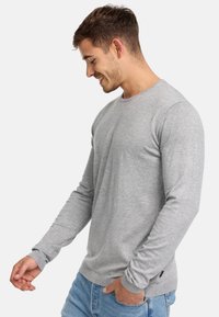 Camiseta de manga larga de color gris claro hecha de tejido suave. Presenta un cuello redondo y mangas ajustadas, combinada con jeans de mezclilla azul.