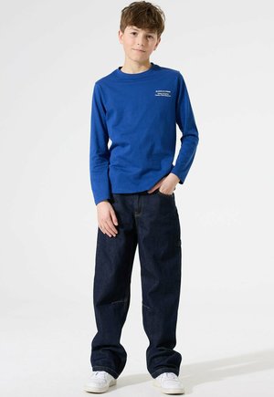 Junge trägt ein blaues Langarmshirt mit Text auf der Brust, lockere dunkelblaue Jeans, weiße Sneakers und steht mit einer Hand in der Tasche.