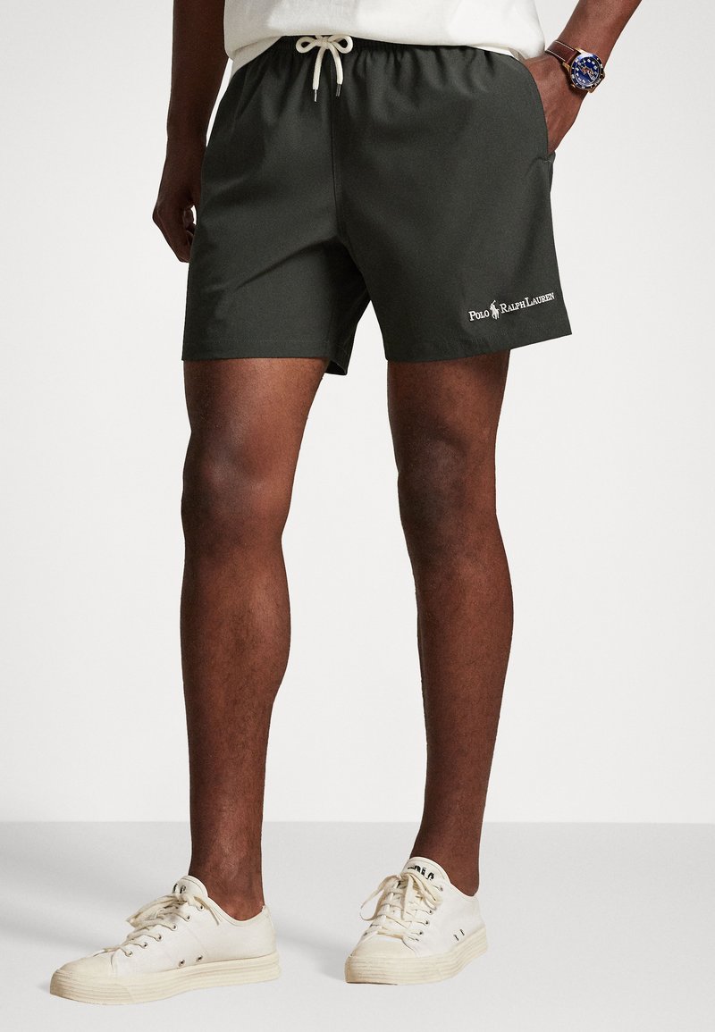 Tmavozelené plavky s elastickým pásom, bielou šnúrkou a vyšitým logom, v kombinácii s bielymi teniskami a každodenným oblečením.