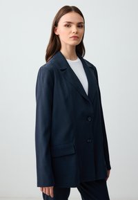 Granatowa marynarka z gładkiego materiału, z klapkami, trzema guzikami, dwoma kieszeniami z przodu i luźnym krojem.