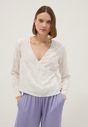 Donna con capelli corti castani che indossa una camicetta bianca a fiori con incrocio e pantaloni larghi lavanda, in piedi con le mani in tasca davanti a uno sfondo semplice.