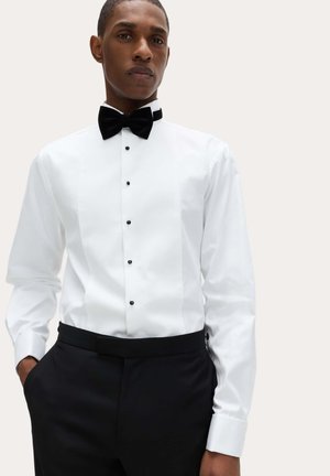 Mężczyzna ubrany w białą koszulę z czarnymi guzikami, czarną muszkę i czarne eleganckie spodnie, stojący z rękami w kieszeniach.