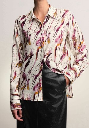 Blouse blanche à manches longues avec des motifs abstraits violets et jaunes portée avec une jupe noire brillante, main dans la poche de la jupe.