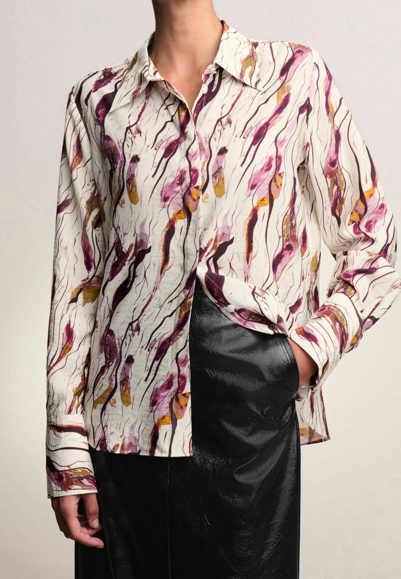 Blouse blanche à manches longues avec des motifs abstraits violets et jaunes portée avec une jupe noire brillante, main dans la poche de la jupe.