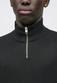 Kotni posnetek osebe, ki nosi črn pleteni pulover s visokim ovratnikom in srebrnim polovičnim zadrgo ter majhno zlato uhano na levi ušesi.