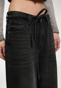 Zbliżenie na osobę noszącą wyblakłe czarne dżinsy, z zawiązanym paskiem i skróconą szaro-długą koszulą, odsłaniającą brzuch.