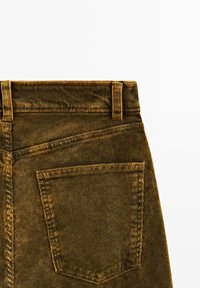 Pantalons en velours côtelé marron avec une texture à côtes verticales, dotés d'une poche arrière et de passants de ceinture. Le tissu est doux, créant un design décontracté.