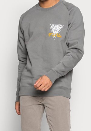 Mann trägt grauen Sweatshirt mit dreieckigem Logo, das einen Tigerkopf zeigt, „DENHAM“-Schriftzug und gelbe japanische Zeichen, kombiniert mit hellbraunen Hosen.