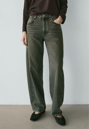 Person iført falmede sorte vidde jeans, en mørkstribet top, sorte flade sko, stående med den ene hånd i lommen mod en ensfarvet baggrund.