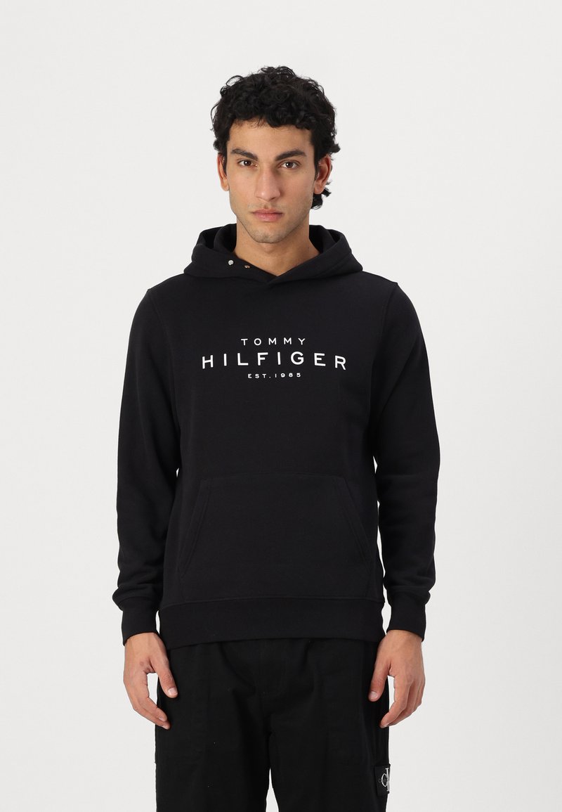 Svart bomullshuvtröja med framficka, snörning i huvan och vit "TOMMY HILFIGER EST. 1985" logotyp på bröstet. Enkel design.