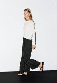 Wit longsleeve top, zwarte wijde broek en bruine slippers. Model staat op een zwarte vloer tegen een effen witte achtergrond.