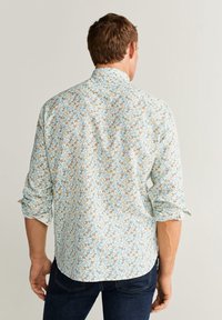 Mann in hellblauem, blumigen Langarmhemd mit hochgekrempelten Ärmeln, kombiniert mit dunkelblauen Jeans, mit dem Rücken zur Kamera gewandt.