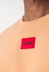 Jasnopomarańczowa bluza z prążkowanym dekoltem. Posiada czerwony prostokątny naszywkę z napisem "HUGO" w kolorze czarnym. Miękka, teksturowana tkanina.