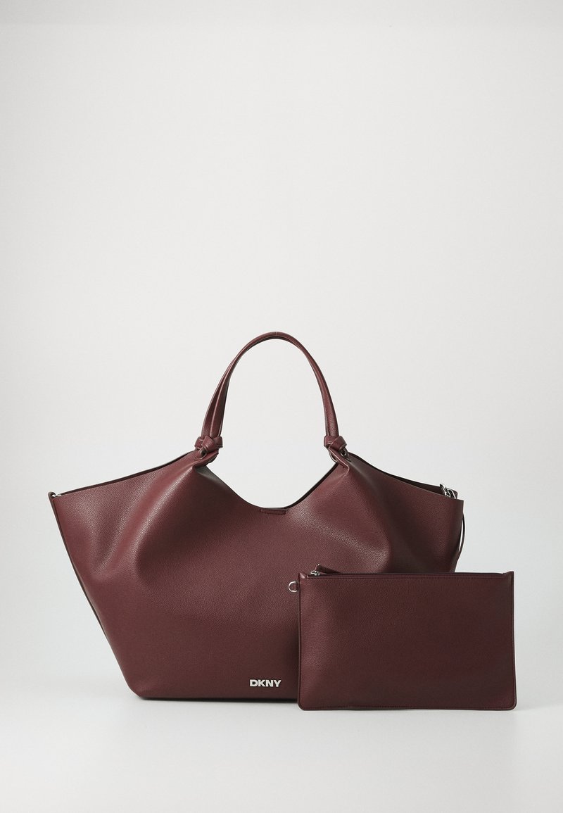 Dark burgundy leather tote bag with a rounded top, featuring dual handles and a detachable zippered pouch. Embossed "DKNY" logo on front.