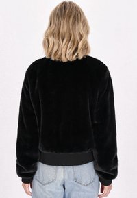 Jachetă din blană artificială neagră cu o croială lejeră, cu manșete și tiv striate. Textură moale cu un guler rotund și cusături vizibile pe spate.