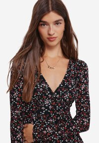 Svart klänning med ett blommönster i vitt, rött och rosa. Har en djup V-ringning och långa ärmar. Guldhalsband med hjärtdesign.