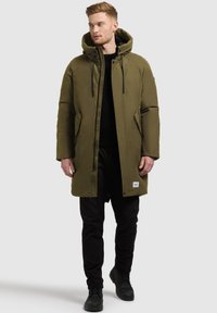 Olivza zaļa puffer jaka ar kapuci, aprīkota ar sānu kabatām, priekšējo rāvējslēdzēju un melniem aukliņām. Valkāta virs melna džempera un biksēm.