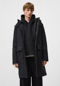 Czarny, długi, ocieplany płaszcz z kapturem, z zamkiem błyskawicznym i zatrzaskami, dwoma przednimi kieszeniami oraz gładkim, teksturowanym materiałem. Nosi się go na szarym hoodie.