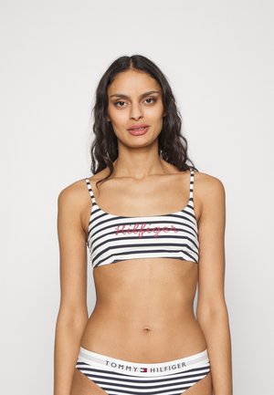 Set bikini a righe in bianco e nero con spalline sottili. La parte superiore presenta il logo rosso "Hilfiger", mentre la parte inferiore mostra "TOMMY HILFIGER" sull'elastico in vita.