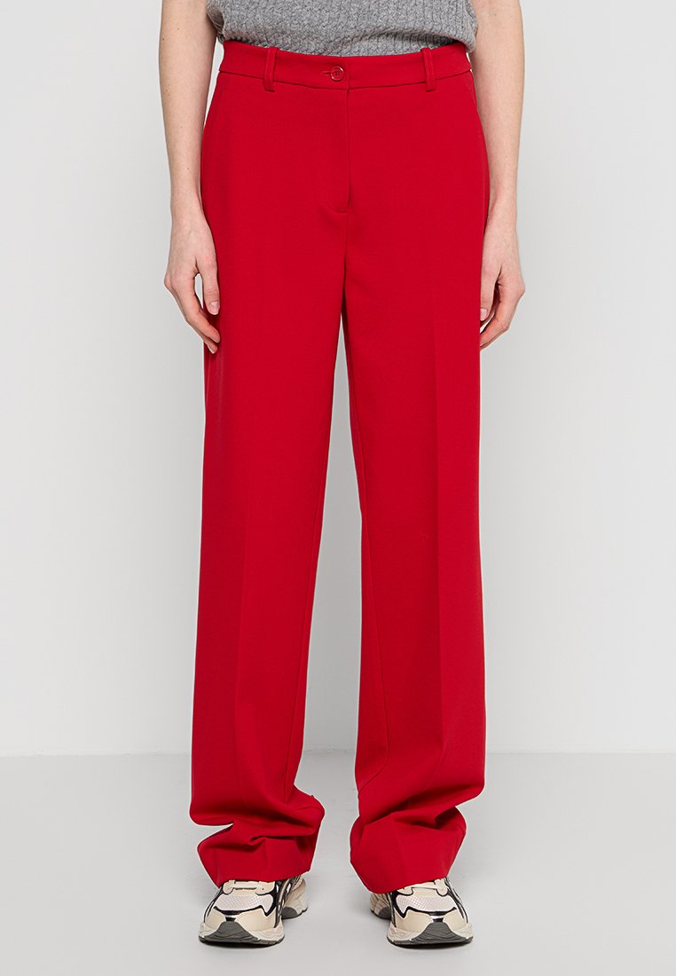 Persona che indossa pantaloni rossi larghi e sneakers bianche con dettagli neri e beige, in piedi davanti a uno sfondo chiaro e semplice.