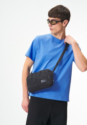 Junger Mann mit schwarzen Sonnenbrillen trägt ein blaues T-Shirt und eine schwarze Umhängetasche, er stellt den Riemen der Tasche vor einem schlichten weißen Hintergrund ein.