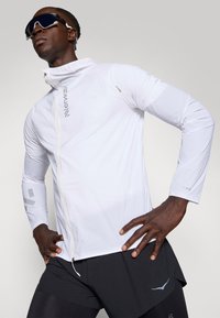 Giacca atletica leggera bianca con chiusura con zip, maniche lunghe, tessuto testurizzato, dettagli riflettenti, abbinata a pantaloni corti neri.
