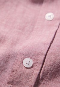 Lichtroze linnen overhemd met een gladde textuur, voorzien van een witte knop en zichtbare stiksels. Close-up van de stof en het knoopontwerp.