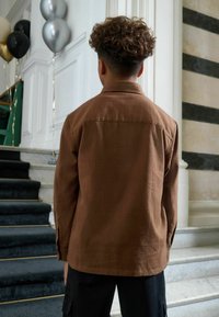 Bruine textuur shirt met een kraag en lange mouwen, gedragen over zwarte cargobroek. Op de achtergrond is een trap en ballonnen te zien.