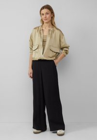 Metallisch goldene Bomberjacke mit zwei Fronttaschen, engen Bündchen und Reißverschluss, kombiniert mit schwarzen weiten Hosen und cremefarbenen Sneakers.