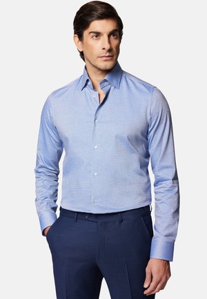 Jasnoniebieska koszula z długim rękawem, o subtelnym wzorzystym wykończeniu, z kontrastującymi detalami mankietów, noszona z ciemnogranatowymi spodniami.