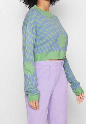 Personne portant un pull court à motifs géométriques verts et violets et un pantalon taille haute lavande sur un fond clair.