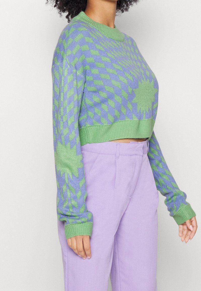 Personne portant un pull court à motifs géométriques verts et violets et un pantalon taille haute lavande sur un fond clair.