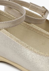 Guldglitrende balletflade med en glat ydre overflade, ankelrem og en blød beige indvendig foring. Har metalliske detaljer langs kanten.