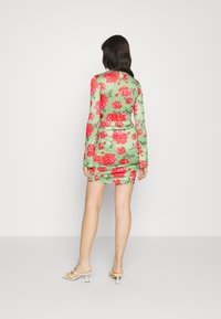 O vestido mini com estampa floral apresenta um tecido verde com grandes rosas vermelhas, mangas longas, um design franzido e uma silhueta ajustada.