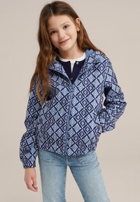 Blå mönstrad zip-up jacka med diamantformer, elastiska manschetter och huva, som är kombinerad med ljusblå jeans.