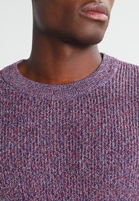 Pull-over en tricot dans un mélange de teintes violettes et rouges, présentant un col arrondi et un tissu texturé avec un motif à mailles bouclées.
