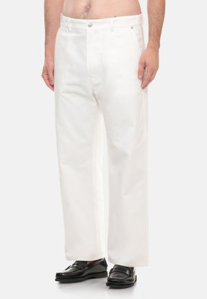 Uomo con pantaloni bianchi a gamba dritta e mocassini neri in pelle, in piedi davanti a uno sfondo semplice.