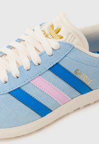 Baskets en daim bleu clair avec trois rayures en bleu et rose, texte "Gazelle" en doré et lacets crème. Semelle en caoutchouc blanche texturée.