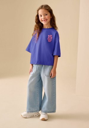 Lila túlméretezett póló rózsaszín virágmintával a mellkason, világoskék bő szárú farmerrel és fehér tornacipővel párosítva.