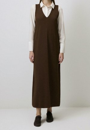 Femme portant une longue robe marron sans manches par-dessus une chemise blanche à manches longues avec col, associée à des baskets foncées, debout devant un fond uni.