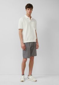 Witte gebreide poloshirt met een zak, gecombineerd met een grijze short. Model draagt witte en zwarte sneakers en staat tegen een lichte achtergrond.